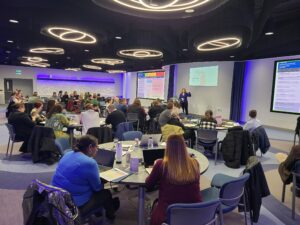 A speaker addresses an audience seated at tables.